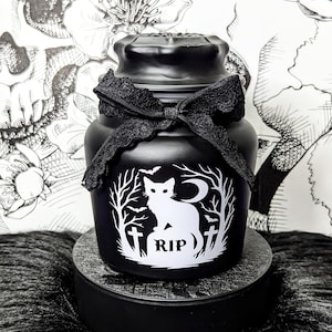 Może przedstawiać: Czarny słoik z dekoracyjną czarną kokardą. Słoik posiada białą grafikę kota, drzew, półksiężyca i napis "RIP". Słoik stoi na czarnej platformie.