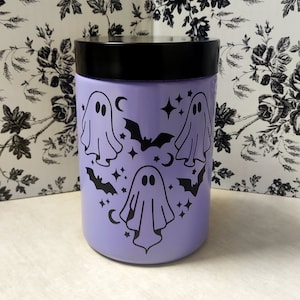 Puede incluir: Un tarro de color lavanda con tapa negra presenta un diseño de tres fantasmas y murciélagos. Los fantasmas son blancos y los murciélagos son negros. El tarro está decorado con estrellas y medias lunas.