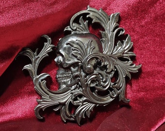 May include: A silver skull with ornate filigree details, set against a deep red velvet background.