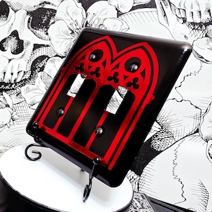 May include: A black light switch cover plate featuring a red gothic arch design with two rectangular cutouts for switches and two black screws. The plate is displayed on a small black metal stand, showcasing a bold contrast between the red and black.