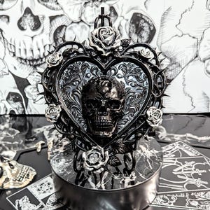 May include: A black, gothic-style heart-shaped decorative piece featuring a skull at its center, surrounded by roses and thorns. The piece sits atop a round, marble-patterned base, with tarot cards and a skull in the background.