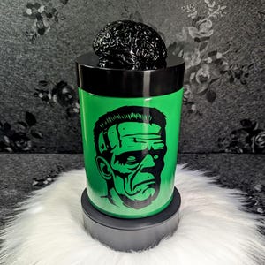 Op de afbeelding: Een groene container met een zwart deksel en een zwart hersenvormig opzetstuk. De container heeft een zwart-groene afbeelding van Frankenstein's monster. De container staat op een zwarte basis en een wit, pluizig oppervlak.
