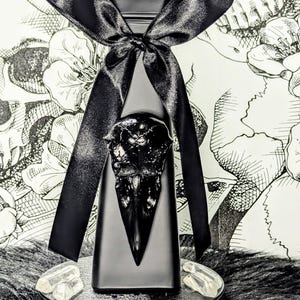Op de afbeelding: Een zwarte vaas met een raven schedel ontwerp, versierd met een zwart satijnen lint. De vaas bevat groene bladeren en staat op een ronde zwarte standaard met heldere kristallen. De achtergrond toont een bloemenpatroon.