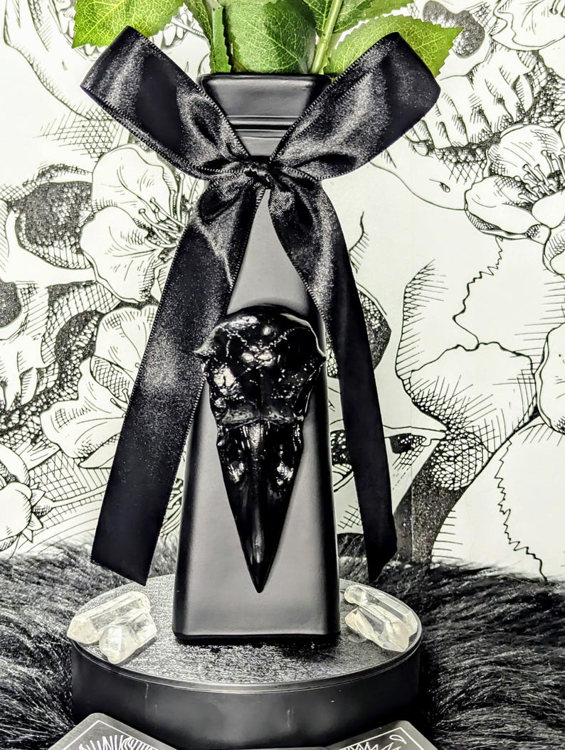Op de afbeelding: Een zwarte vaas met een raven schedel ontwerp, versierd met een zwart satijnen lint. De vaas bevat groene bladeren en staat op een ronde zwarte standaard met heldere kristallen. De achtergrond toont een bloemenpatroon.