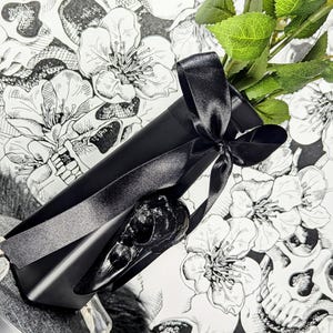 Op de afbeelding: Een zwarte geschenkdoos met een zwart satijnen lint, met een zwarte raven schedel. De doos ligt op een bloemen- en schedelpatroon achtergrond, met groene bladeren. Een gothic cadeau.