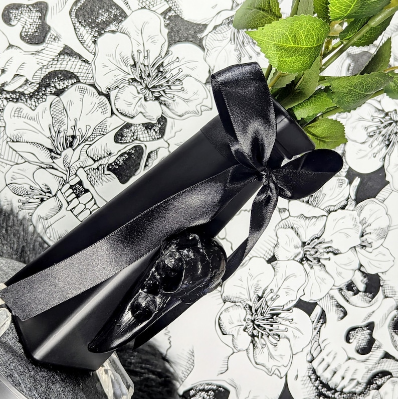 Op de afbeelding: Een zwarte geschenkdoos met een zwart satijnen lint, met een zwarte raven schedel. De doos ligt op een bloemen- en schedelpatroon achtergrond, met groene bladeren. Een gothic cadeau.