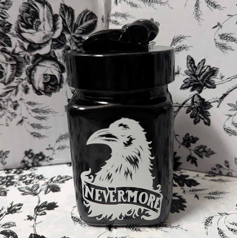 May include: Black glass jar with a black skull-shaped lid. The jar has a white raven design and the words "NEVERMORE" printed on it.