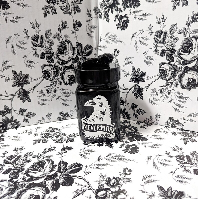 May include: A black glass bottle with a black screw top lid. The bottle has a white raven graphic with the words 'NEVERMORE' printed below the raven.