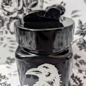 May include: Black glass jar with a black skull lid and a white raven design. The jar has a distressed, aged look.