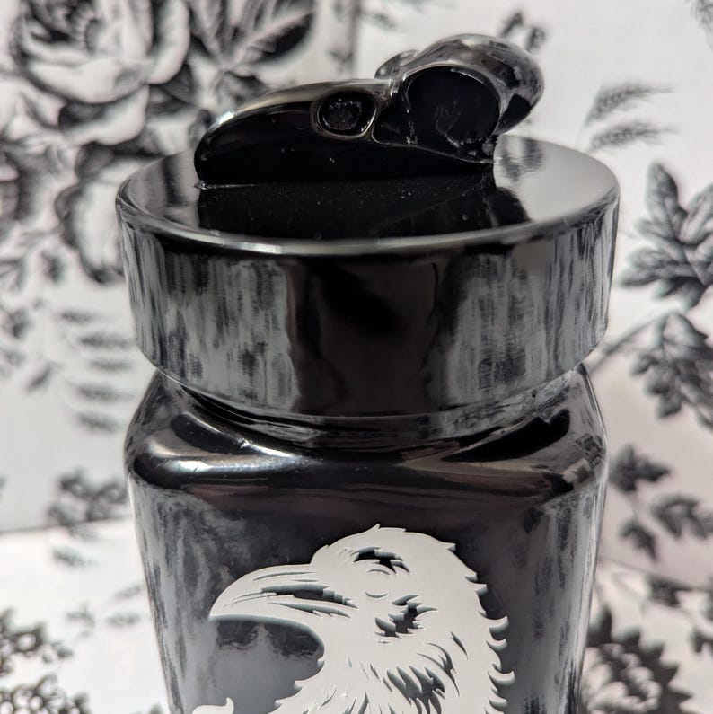 May include: Black glass jar with a black skull lid and a white raven design. The jar has a distressed, aged look.