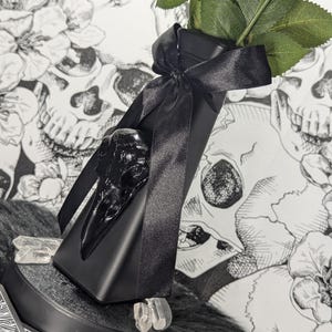 Op de afbeelding: Een zwarte decoratieve vaas met een raven schedel accent en een zwart satijnen lint. De vaas bevat een stengel met groene bladeren. Heldere kristallen zijn rond de basis geplaatst. De achtergrond heeft een zwart-wit schedel- en bloemenpatroon.