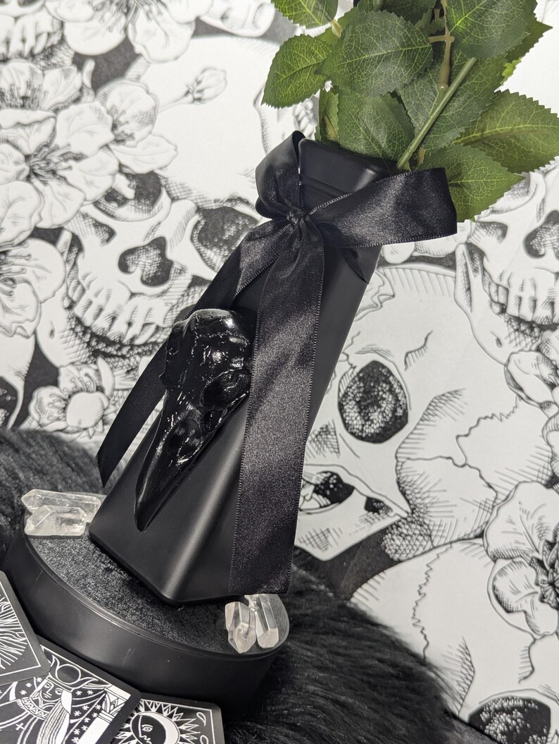 Op de afbeelding: Een zwarte decoratieve vaas met een raven schedel accent en een zwart satijnen lint. De vaas bevat een stengel met groene bladeren. Heldere kristallen zijn rond de basis geplaatst. De achtergrond heeft een zwart-wit schedel- en bloemenpatroon.
