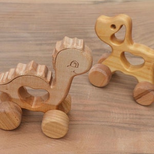 Può includere: Due giocattoli di legno da tirare, un dinosauro e un cane, entrambi con ruote. I giocattoli sono realizzati in legno naturale e hanno un colore marrone chiaro.