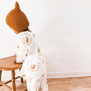 Könnte beinhalten: Ein Kind trägt eine braune Strickmütze mit Katzenohren und ein weißes Langarmshirt mit braunem Sonnenmuster. Das Kind steht neben einem Holzhocker.