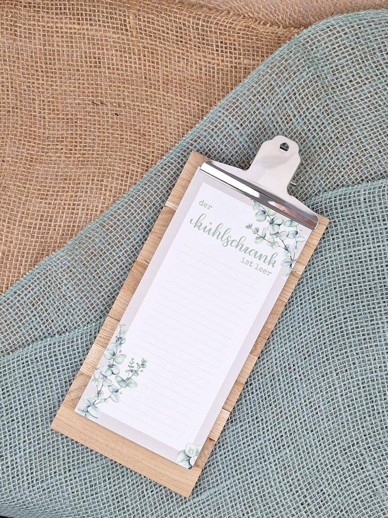 Puede incluir: Un portapapeles rectangular de madera con un bloc de notas rayado y un clip de metal plateado. El bloc de notas tiene un fondo blanco con ilustraciones de hojas de eucalipto verdes y el texto en alem&aacute;n "der k&uuml;hlschrank ist leer".