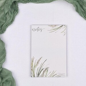 Puede incluir: Un bloc de notas rectangular con fondo blanco que presenta la palabra notes en letra cursiva en la parte superior izquierda. El bloc de notas está decorado con ilustraciones en acuarela de hierbas verdes y beige en las esquinas superior derecha e inferior izquierda.