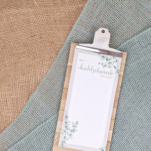 Puede incluir: Un portapapeles de madera con un bloc de notas rayado y un clip de metal plateado. El bloc de notas tiene un fondo blanco con ilustraciones de hojas de eucalipto verdes y el texto en alem&aacute;n "der k&uuml;hlschrank ist leer" en letra cursiva.