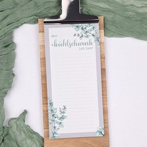 Puede incluir: Un portapapeles de madera que sostiene un bloc de notas vertical con un borde gris e ilustraciones de hojas de eucalipto. El bloc presenta el texto "der k&uuml;hlschrank ist leer" en la parte superior e incluye l&iacute;neas horizontales para escribir.