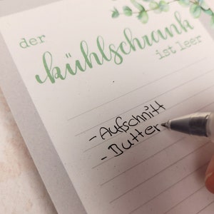 Puede incluir: Un bloc de notas blanco con l&iacute;neas horizontales gris claro y una ilustraci&oacute;n de hojas de eucalipto verdes en la parte superior. El texto "der k&uuml;hlschrank ist leer" est&aacute; impreso en verde. Un bol&iacute;grafo negro escribe "Aufschnitt" y "Butter".