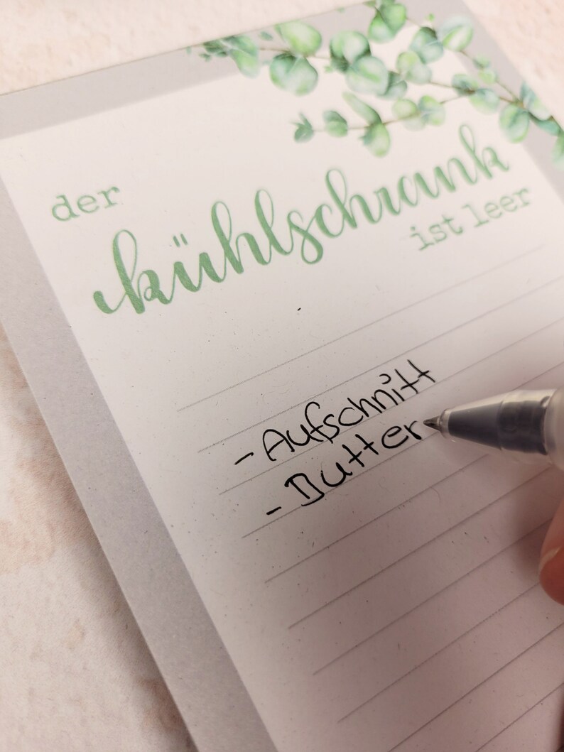 Puede incluir: Un bloc de notas blanco con l&iacute;neas horizontales gris claro y una ilustraci&oacute;n de hojas de eucalipto verdes en la parte superior. El texto "der k&uuml;hlschrank ist leer" est&aacute; impreso en verde. Un bol&iacute;grafo negro escribe "Aufschnitt" y "Butter".