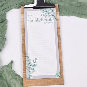 Puede incluir: Un portapapeles rectangular de madera que sostiene un bloc de notas rayado con un borde gris e ilustraciones de hojas de eucalipto verdes. El bloc de notas presenta el texto impreso en alem&aacute;n "der k&uuml;hlschrank ist leer" y un clip de metal plateado.