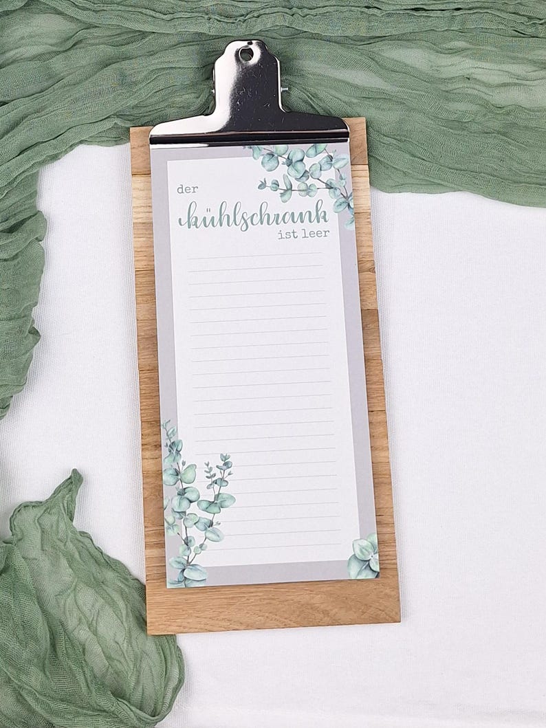 Puede incluir: Un portapapeles rectangular de madera que sostiene un bloc de notas rayado con un borde gris e ilustraciones de hojas de eucalipto verdes. El bloc de notas presenta el texto impreso en alem&aacute;n "der k&uuml;hlschrank ist leer" y un clip de metal plateado.