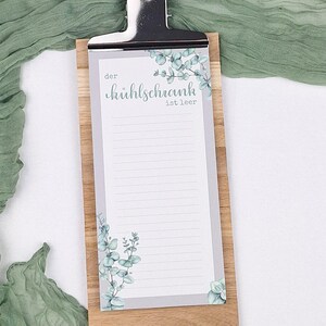 Puede incluir: Un portapapeles de madera que sostiene un bloc de notas rayado con un borde gris e ilustraciones de hojas de eucalipto verdes. El bloc de notas presenta el texto en alem&aacute;n "der k&uuml;hlschrank ist leer" impreso en una fuente cursiva.