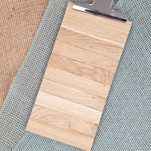 Puede incluir: Un portapapeles rectangular construido con m&uacute;ltiples listones de madera de color claro dispuestos horizontalmente. La parte superior cuenta con un mecanismo de clip de metal plateado pulido con un peque&ntilde;o orificio circular.