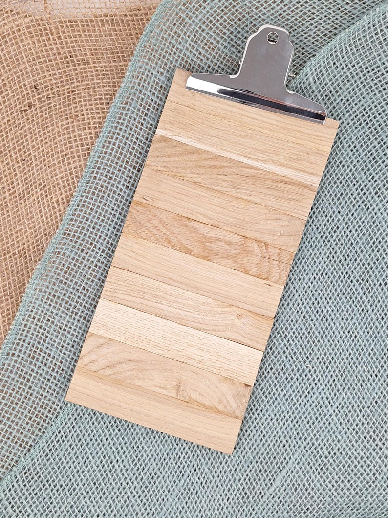Puede incluir: Un portapapeles rectangular construido con m&uacute;ltiples listones de madera de color claro dispuestos horizontalmente. La parte superior cuenta con un mecanismo de clip de metal plateado pulido con un peque&ntilde;o orificio circular.