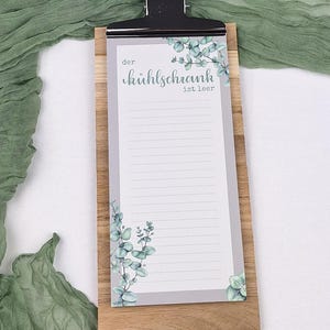 Puede incluir: Un bloc de notas rectangular con un clip de metal negro sujeto a una tabla de madera. El papel presenta un fondo gris claro con ilustraciones de hojas de eucalipto verdes en la parte superior e inferior, y el texto "der k&uuml;hlschrank ist leer".