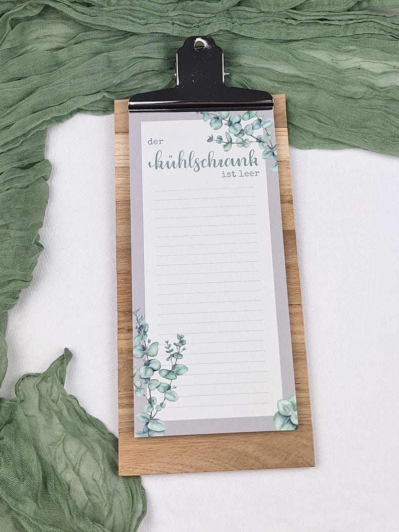Puede incluir: Un bloc de notas rectangular con un clip de metal negro sujeto a una tabla de madera. El papel presenta un fondo gris claro con ilustraciones de hojas de eucalipto verdes en la parte superior e inferior, y el texto "der k&uuml;hlschrank ist leer".
