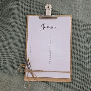 Puede incluir: Un portapapeles de madera con un calendario de papel blanco para el mes de enero. El calendario tiene un diseño simple con los días del mes enumerados en una columna a la izquierda y las fechas correspondientes a la derecha. El portapapeles está decorado con un trozo de cuerda y flores secas.