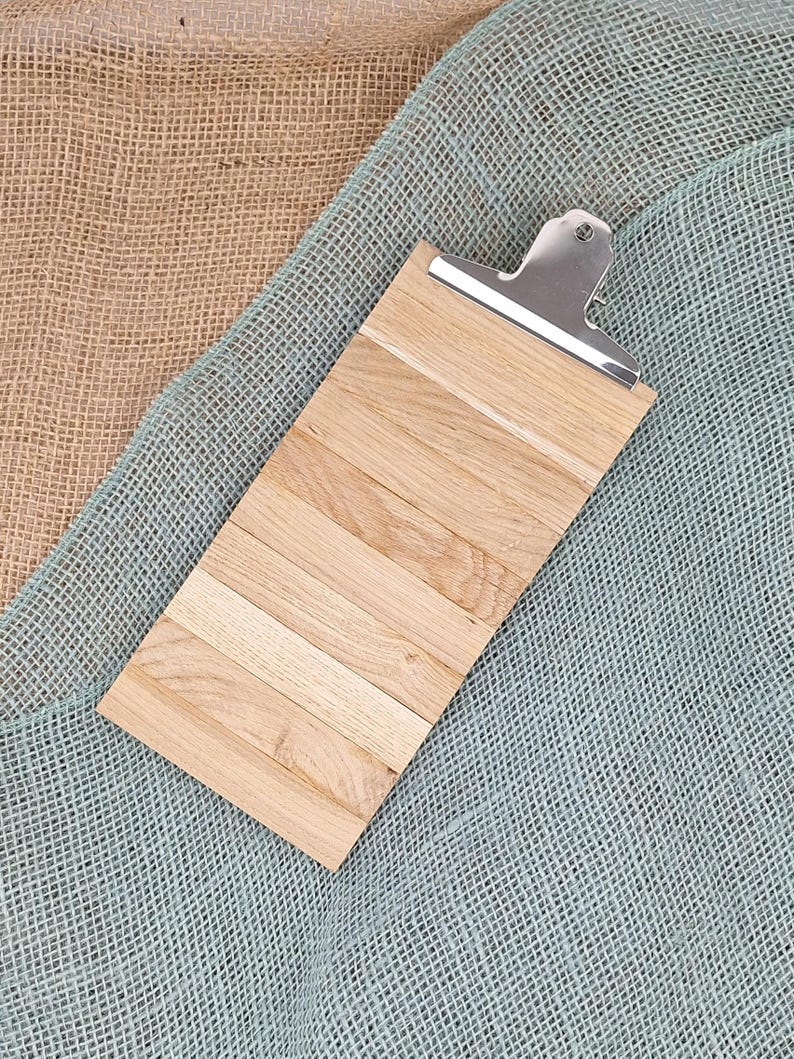 Puede incluir: Un portapapeles rectangular construido con varias l&aacute;minas de madera de color marr&oacute;n claro dispuestas horizontalmente una al lado de la otra. La parte superior cuenta con un mecanismo de clip de metal plateado pulido.
