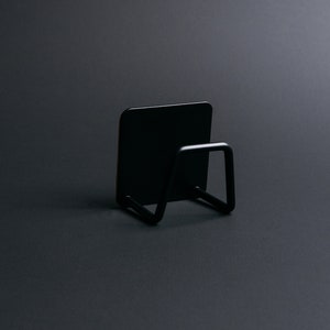 Può includere: Un supporto minimalista in metallo nero con schienale quadrato e maniglia ad anello. Il supporto è progettato per contenere un piccolo oggetto, come un blocco note o un telefono. L'oggetto è posto su uno sfondo grigio scuro.