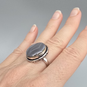 Könnte beinhalten: Ein Silberring mit einem großen, ovalen, grauen Edelstein mit einem helleren grauen Streifen. Der Edelstein ist in einer silbernen Lünette mit dekorativem Rand gefasst. Der Ring befindet sich an einem Finger.