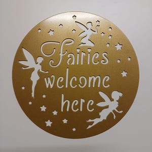 May include: A gold round sign with the words "Fairies welcome here" cut out in white. Three white fairy silhouettes are also cut out on the sign.