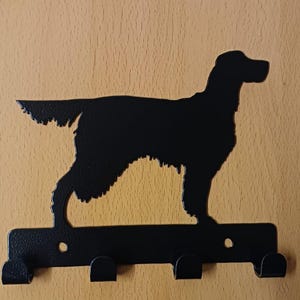 May include: Black metal wall-mounted key holder featuring a silhouette of a dog. The key holder has three hooks below the dog's image. The dog is in a standing position with its tail up. The key holder is mounted on a light brown wooden surface.