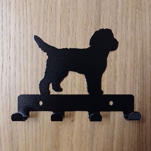 May include: Black metal key hook with a silhouette of a dog. The hook has four hooks and is mounted on a wooden surface.