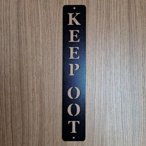 Peut inclure: Panneau en métal noir avec les mots « KEEP OUT » découpés. Le panneau est rectangulaire avec un petit trou en haut pour l'accrocher. Le panneau est affiché sur un fond en bois. Les mesures sont en centimètres.