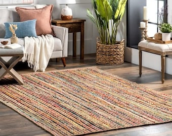 May include: A multi-colored striped jute rug with a natural fiber texture. The rug is laid out on a hardwood floor in a living room setting with a sofa, a side table, a plant, and a bench.