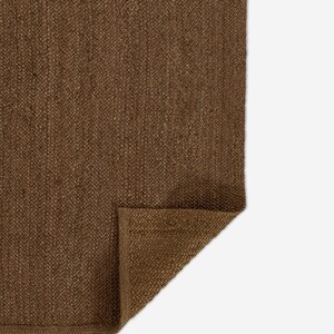 Puede incluir: Una alfombra rectangular de color marr&oacute;n con una textura tejida densa y un patr&oacute;n trenzado visible. Una esquina de la alfombra est&aacute; doblada, revelando la parte inferior del material y un borde recto acabado a lo largo del per&iacute;metro.
