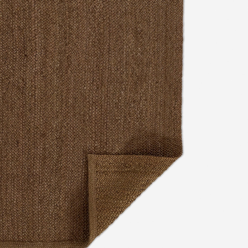Puede incluir: Una alfombra rectangular de color marr&oacute;n con una textura tejida densa y un patr&oacute;n trenzado visible. Una esquina de la alfombra est&aacute; doblada, revelando la parte inferior del material y un borde recto acabado a lo largo del per&iacute;metro.