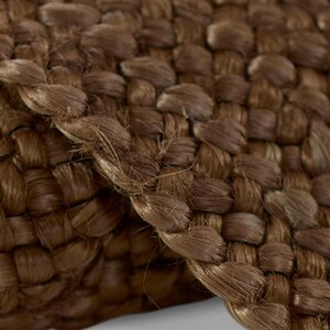Puede incluir: Un primer plano de una alfombra tejida de color marr&oacute;n con una textura trenzada gruesa y voluminosa. El material muestra un aspecto natural y fibroso con hebras visibles y un patr&oacute;n denso entrelazado que crea una superficie t&aacute;ctil.