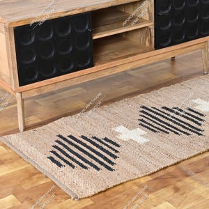 May include: A rectangular jute runner rug featuring a natural tan base with repeating black geometric striped patterns and white cross-shaped accents. The rug is positioned on a wooden floor in front of a wooden media console with black doors.