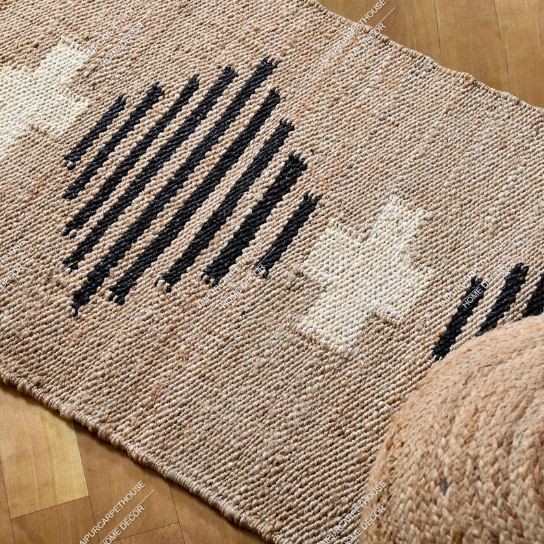 May include: A rectangular woven rug made of natural tan fibres featuring a repeating pattern of black diagonal stripes and white cross shapes. The rug has a textured, braided appearance with a consistent weave throughout the entire surface area.