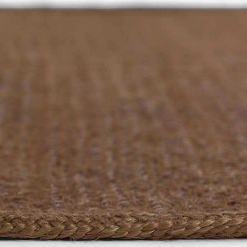Puede incluir: Primer plano de una alfombra tejida marr&oacute;n con un borde trenzado. La alfombra tiene una superficie texturizada y un tono natural y terroso. El patr&oacute;n de tejido es visible, creando un dise&ntilde;o sutil. La alfombra parece estar hecha de fibras naturales.