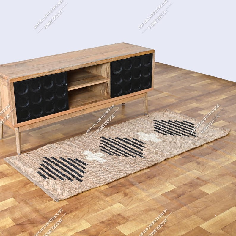 May include: A rectangular jute runner rug featuring a natural tan base with a repeating geometric pattern of black stripes and white cross shapes. The rug is positioned on a wooden floor in front of a wooden media console with two black doors.