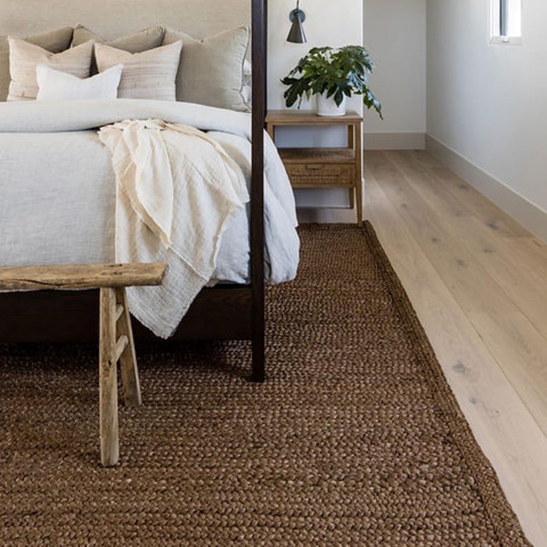 Puede incluir: Una habitaci&oacute;n con una alfombra tejida marr&oacute;n oscuro. La alfombra es rectangular y cubre parte del suelo. Un banco de madera est&aacute; al pie de una cama con un edred&oacute;n y almohadas blancos. Tambi&eacute;n se ven una mesita de noche de madera y una planta en maceta.