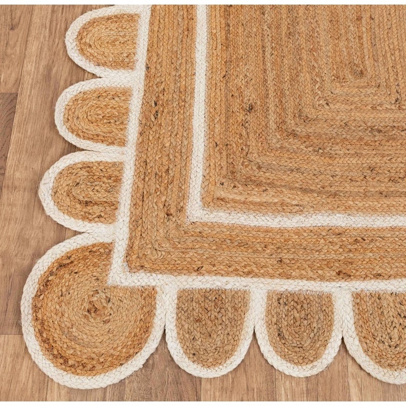 Puede incluir: Una alfombra de fibra natural con forma cuadrada y un borde blanco. La alfombra tiene un borde festoneado con un ribete blanco.