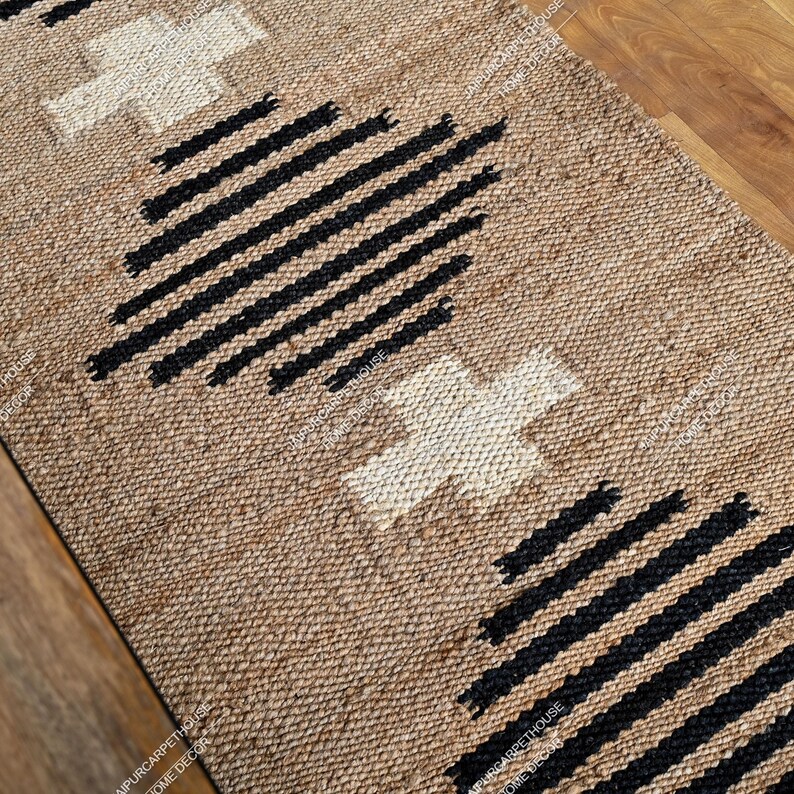May include: A rectangular woven rug made of natural tan fibres featuring a repeating pattern of white cross shapes and black horizontal striped diamond motifs. The textured surface displays a consistent braided weave throughout the entire piece.